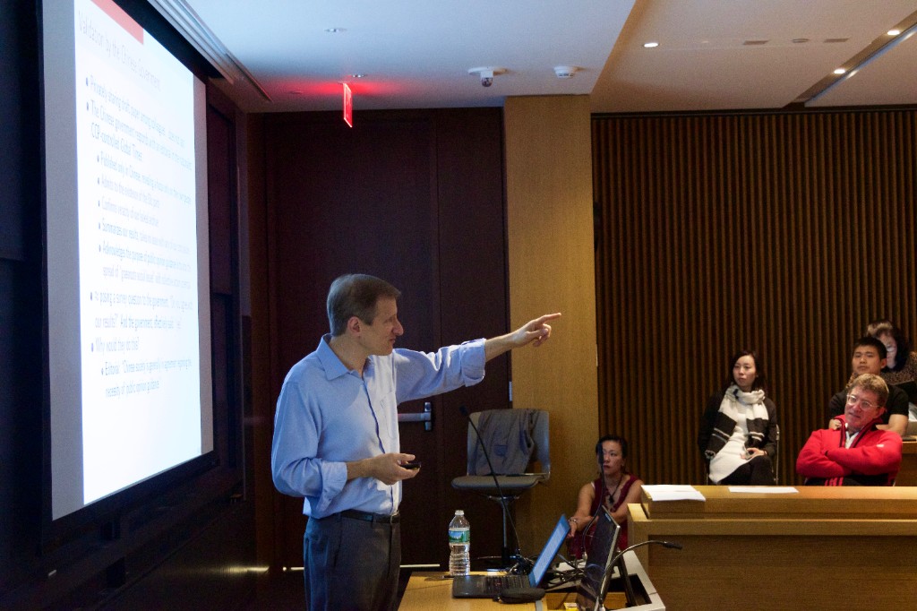 Gary King presenting in a lecture hall: projected slides on a large screen, wooden podium with laptop, tiered seating with audience members.